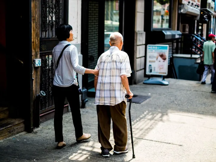 Une femme accompagne un homme âgé avec une canne sur un trottoir.